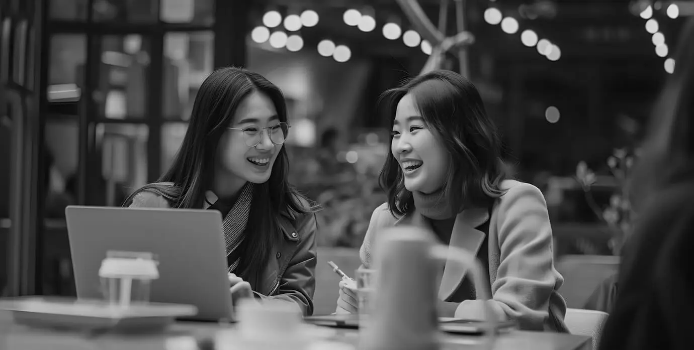 Black and white photo of two happy women smiling and laughing together at a laptop in a cafe.