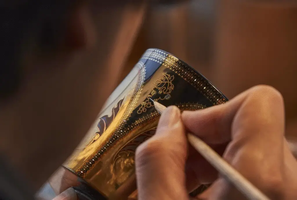 Close-up of an artisan's hand meticulously painting an intricate gold and black decorative design onto a curved object.