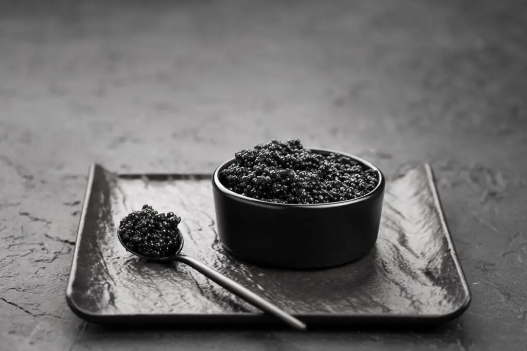 Close-up of a bowl of black caviar with a spoon on a textured plate, presented in black and white.